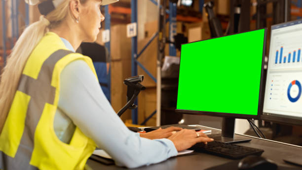 Professional Female Worker Wearing Hard Hat Uses Computer with Green Chroma Screen and Inventory Delivery Software in the Retail Warehouse full of Shelves with Goods. Distribution Logistical Center Professional Female Worker Wearing Hard Hat Uses Computer with Green Chroma Screen and Inventory Delivery Software in the Retail Warehouse full of Shelves with Goods. Distribution Logistical Center logistical stock pictures, royalty-free photos & images