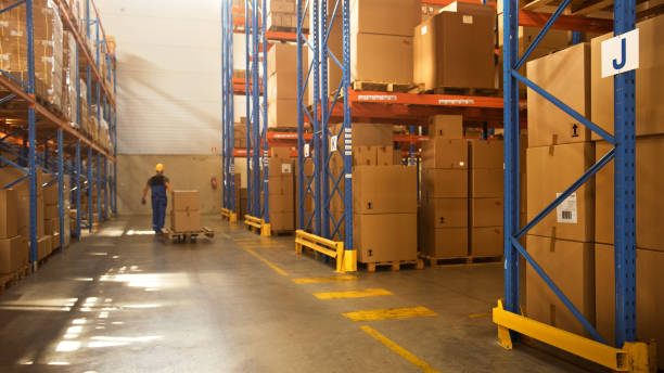 istock Worker Moves Cardboard Boxes using Manual Pallet Truck, Walking between Rows of Shelves with Goods in Retail Warehouse. People Work in Product Distribution Logistics Center. Side View 1283514249