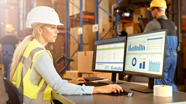 istock Professional Female Worker Wearing Hard Hat Uses Computer with Inventory Status Checking and Delivery Software in the Retail Warehouse full of Shelves with Goods. Delivery, Distribution Center 1283514303
