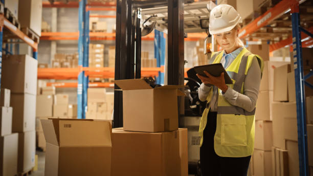 In Warehouse Female Manager Wearing Hardhat Standing beside Forklift Uses Digital Tablet for Inventory, Checking Inside of Cardboard Box Package. Distribution, Delivery Center, Shelves with Packages en almacén gerente femenino usando hardhat standing al lado de carretilla elevadora utiliza tableta digital para el inventario, comprobando el interior del paquete de la caja de cartón. distribución, centro de entrega, estantes con paquetes