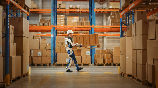 High-Tech Futuristic Warehouse: Manager Scans Packages for Inventory, Delivery in the Background Worker Wearing Advanced Full Body Powered exoskeleton, Walks with Heavy Pallet full of Cardboard Boxes almacén futurista de alta tecnología: el gerente escanea los paquetes para el inventario, la entrega en el trabajador de fondo que lleva el exoesqueleto avanzado alimentado por todo el cuerpo, camina con el palé pesado lleno de cajas de cartón