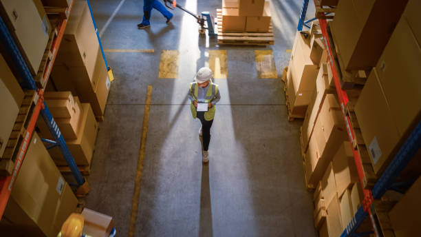 istock Top-Down View: Worker Wearing Hard Hat Checks Stock and Inventory Using Digital Tablet Computer in the Retail Warehouse full of Shelves with Goods. Working in Logistics, Distribution 1283514451