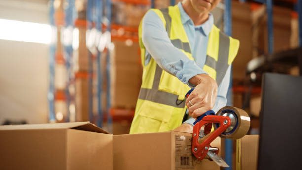 istock In Retail Warehouse Professional Worker Packing Parcel, Cardboard Box Sealed with Tape Dispenser Ready for Shipment. Delivery and Distribution Center Full of Shelves with Goods. Close-up Shot 1283514482