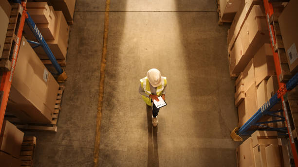 Top-Down View: Worker Wearing Hard Hat Checks Stock and Inventory Using Digital Tablet Computer in the Retail Warehouse full of Shelves with Goods. Working in Logistics, Distribution Top-Down View: Worker Wearing Hard Hat Checks Stock and Inventory Using Digital Tablet Computer in the Retail Warehouse full of Shelves with Goods. Working in Logistics, Distribution shipping stock pictures, royalty-free photos & images