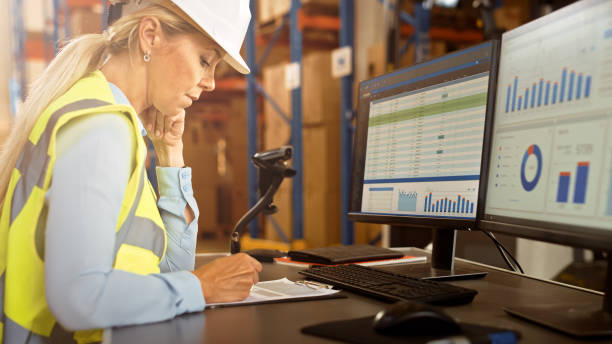 istock Professional Female Worker Wearing Hard Hat Uses Computer with Inventory Status Checking and Delivery Software in the Retail Warehouse full of Shelves with Goods. Delivery, Distribution Center 1283514495