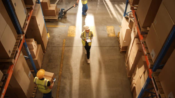 istock Top-Down View: Worker Wearing Hard Hat Checks Stock and Inventory Using Digital Tablet Computer in the Retail Warehouse full of Shelves with Goods. Working in Logistics, Distribution 1283514511