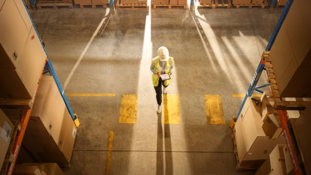 istock Top-Down View: Worker Wearing Hard Hat Checks Stock and Inventory Using Digital Tablet Computer in the Retail Warehouse full of Shelves with Goods. Working in Logistics, Distribution 1283514518