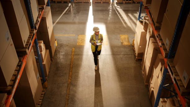 istock Top-Down View: Worker Wearing Hard Hat Checks Stock and Inventory Using Digital Tablet Computer in the Retail Warehouse full of Shelves with Goods. Working in Logistics, Distribution 1283514520