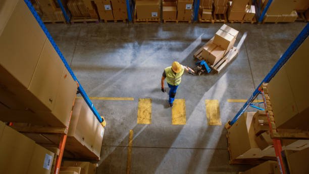 istock Top-Down View: Worker Moves Cardboard Boxes using Manual Pallet Truck, Walking between Rows of Shelves with Goods in Retail Warehouse. People Work in Product Distribution Logistics Center 1283514535