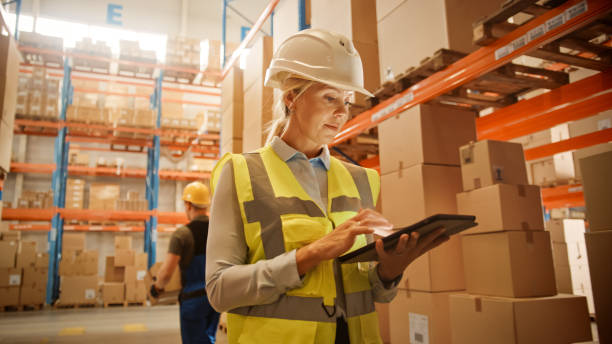 Professional Confident Worker Wearing Hard Hat Checks Stock and Inventory with Digital Tablet Computer in the Retail Warehouse full of Shelves with Goods. Working in Logistics, Distribution Center Professional Confident Worker Wearing Hard Hat Checks Stock and Inventory with Digital Tablet Computer in the Retail Warehouse full of Shelves with Goods. Working in Logistics, Distribution Center logistical stock pictures, royalty-free photos & images