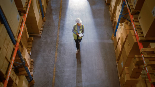 istock Top-Down View: Worker Wearing Hard Hat Checks Stock and Inventory Using Digital Tablet Computer in the Retail Warehouse full of Shelves with Goods. Working in Logistics, Distribution 1283514583