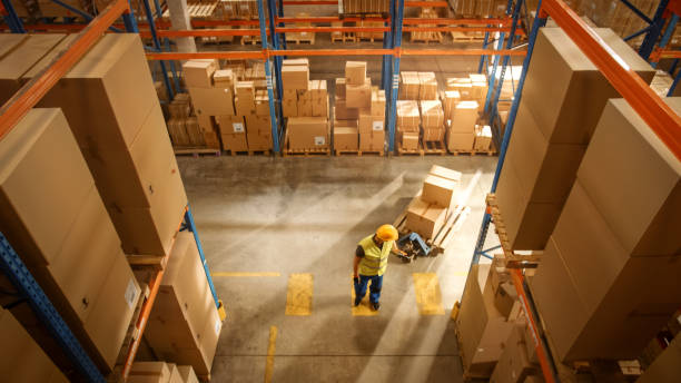 istock Top-Down View: Worker Moves Cardboard Boxes using Manual Pallet Truck, Walking between Rows of Shelves with Goods in Retail Warehouse. People Work in Product Distribution Logistics Center 1283514588