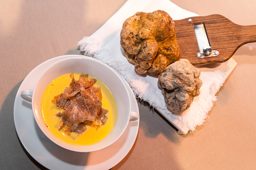 two big white alba truffles on wooden slicer and white napkin near cheese fondue with slices of truffle in