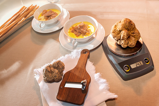 scale weighing a large beautiful white truffle near wooden slicer in the background two cups with cheese fondue and sliced truffles, all on the table