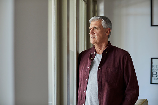 Mature man looking through window. Thoughtful male is at home during COVID-19 lockdown. He is wearing casuals.