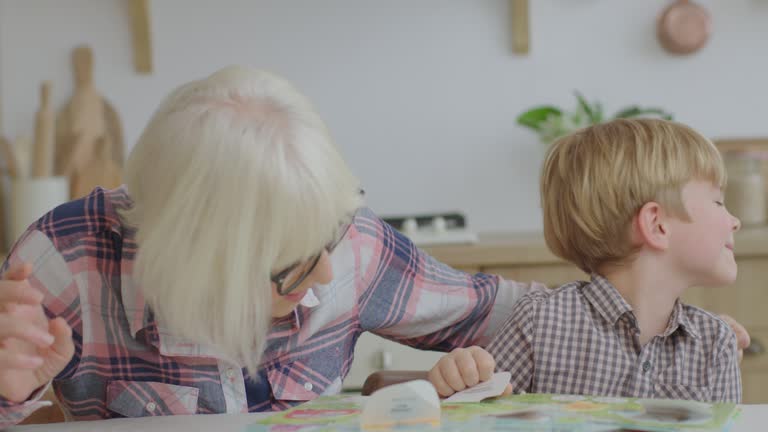 Preschool boy laughing and playing board game with grandmother at wooden kitchen. Happy family reading interesting book together. Granny and grandchild having fun.