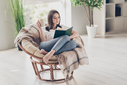 Full size photo of concentrated girl read book sitting on wicker chair with coffee cup wear white t-shirt in house indoors.