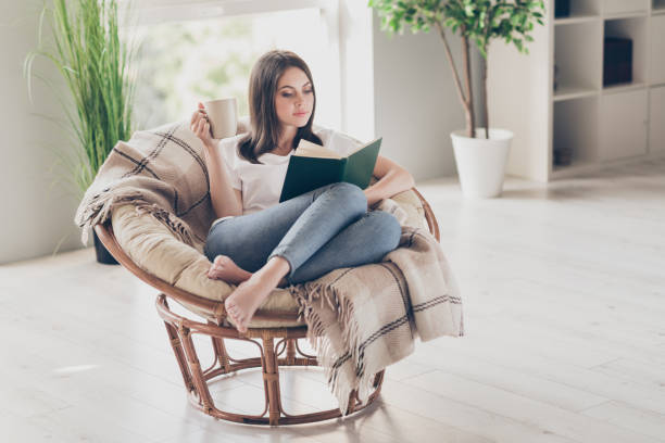 полный размер фото концентрированной девушки читать книги, сидя на плетеном стуле с кофейной чашкой носить белую футболку в доме в помещен� - book reading white women стоковые фото и изображения