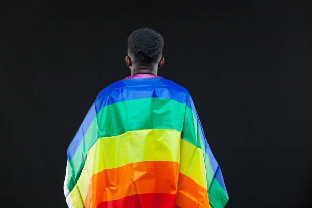 Back view of young african american man wrapped in a rainbow flag isolated on black background. Concept of The LGBT community, minority rights, protection of human rights Back view of young african american man wrapped in a rainbow flag isolated on black background. Concept of The LGBT community, minority rights, protection of human rights. human rights act stock pictures, royalty-free photos & images