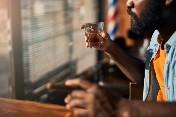 bearded young man smoking cigar and drinking whisky - cigar whisky bar cognac imagens e fotografias de stock