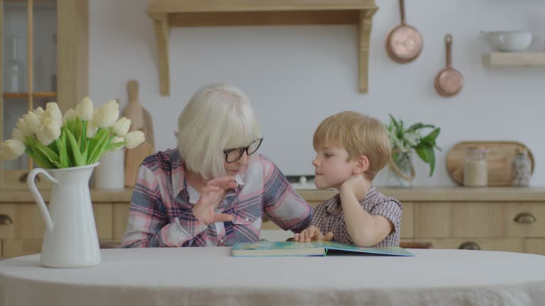 Preschool boy playing board game with grandmother at wooden kitchen. Happy family reading interesting book together. Granny and grandchild having fun.