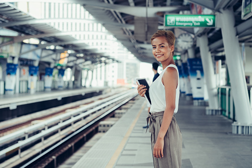 Beautiful short hair blonde female standing at train station, holding digital tablet and looking and smiling at camera