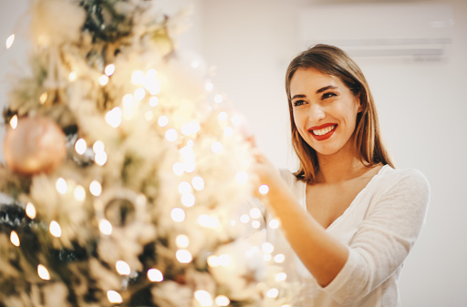 Young woman is fixing decoration on Christmas tree. She is rearranging ornaments, and lights on Christmas tree to make it lighter and sparkler.