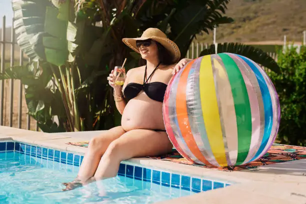 Shot of a pregnant woman sitting by the pool with a drink and a beach ball