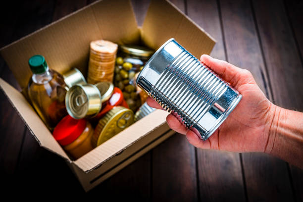 High angle view of a hand picking up a metal tin from non-perishable food cardboard box. The hand holding the tin is in focus while the cardboard box is out of focus. High resolution 42Mp studio digital capture taken with SONY A7rII and Zeiss Batis 40mm F2.0 CF lens