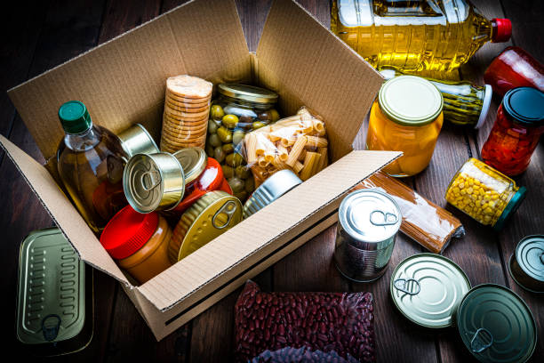 High angle view of a cardboard box filled with multicolored non-perishable canned goods, conserves, sauces and oils shot on wooden table. The composition includes cooking oil bottle, pasta, crackers, preserves and tins. High resolution 42Mp studio digital capture taken with SONY A7rII and Zeiss Batis 40mm F2.0 CF lens
