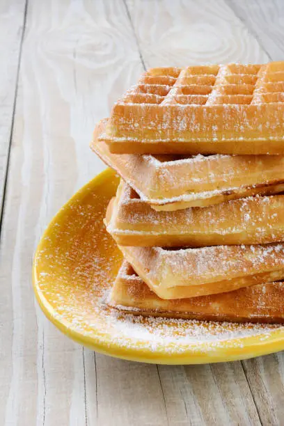 Closeup of a place Belgian Waffles covered with powdered sugar. The stack of waffles is on a yellow platter on a rustic farmhouse style kitchen table.