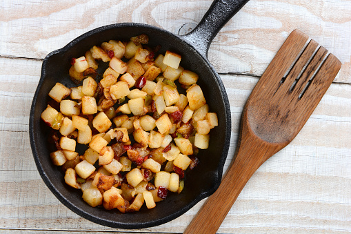 Fried Breakfast Potatoes in a cast iron skillet. Peppers, onions and potato cubes fill the skillet resting on a rustic farmhouse style kitchen table with a wooden fork.