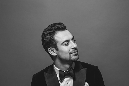 Studio portrait of a young caucasian man in a tuxedo, collar unbuttoned, bow tie untied, looking down, against plain studio background