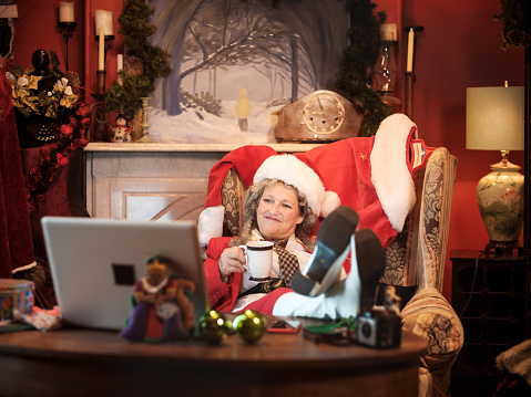 Mrs.Clause using a computer in Santa's study to do a Christmas video conference. She  holding a mug with hot beverage.