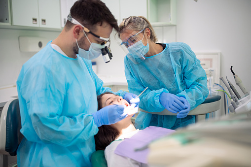 Dentist and her assistant working on young female patient in medical clinic with protective equipment, dental health during covid-19 pandemic.