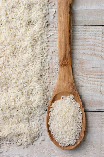 Overhead view of a wooden spoon full of rice next to a pile of more rice on a rustic wood table.