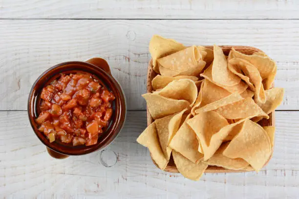 an Overhead view of a bowl of corn chips next ot a pot of salsa on a rustic wood table. Horizontal format.