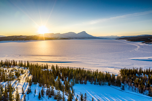 AERIAL: Sunset golden light over Norwegian snowy winter mountains and Rossvatnet lake, pine and birch trees, early spring, calm blue skies. Hattfjelldal municipality, Northern Norway