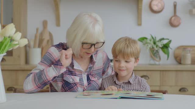 Blonde granny playing board game with preschool grandson at wooden kitchen. Happy senior woman and kid having fun staying at home. Grandmother reads fairytale to grandson.