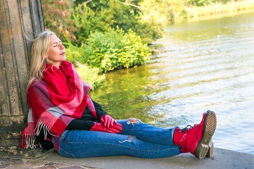 blond woman in jeans and big red scarf sitting outside by a lake
