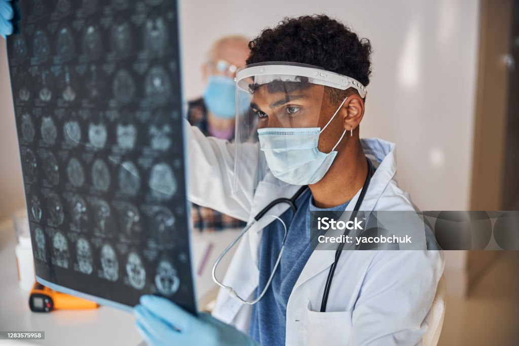 Neurologist staring at the patient brain images Serious young male doctor in a face shield and a mask examining the MRI scans MRI Scan Stock Photo