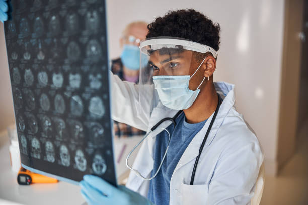 Neurologist staring at the patient brain images Serious young male doctor in a face shield and a mask examining the MRI scans civilian stock pictures, royalty-free photos & images