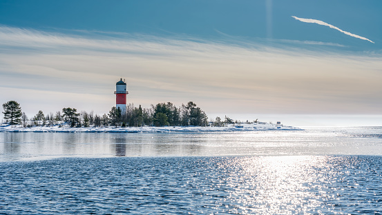Very close view of red white lighthouse in middle of frozen, snowed island at cold Baltic Sea, partly open water, thin ice reflecting day light. Blue sky, light breeze. Northern Sweden, Umea