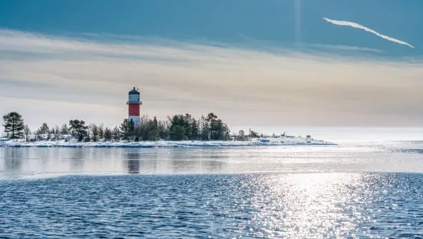 Photo of Very close view of red white lighthouse in middle of frozen, snowed island at cold Baltic Sea, partly open water, thin ice reflecting day light. Blue sky, light breeze. Northern Sweden, Umea