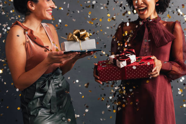 temps de fête : deux femmes élégantes échangeant des cadeaux de noel à une partie, confettis volant tout autour d’eux - black and gold photos et images de collection