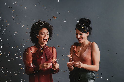 Two happy elegant women holding sparklers and having fun being showered with gold and silver confetti (copy space).