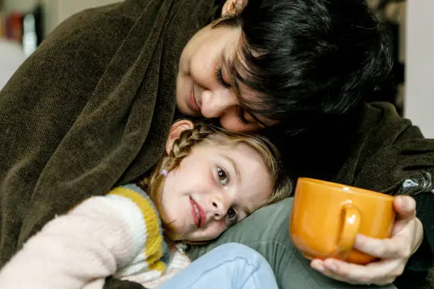 Photo of happy mother with her little daughter at home by the fireplace holding a cup of tea or coffee