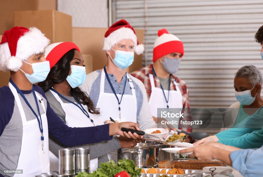 Multi-ethnic group of volunteers work at soup kitchen at Christmas. Multi-ethnic, mixed age group of volunteers work together at food bank, soup kitchen during Christmas season.  They serve food to needy people in their community at holiday times. Face masks. Christmas Stock Photo