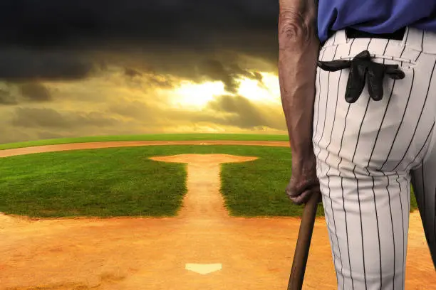 Closeup of a baseball player leaning on his bat seen from behind. Standing on a field looking out to an endless horizon and a dark stormy sky.