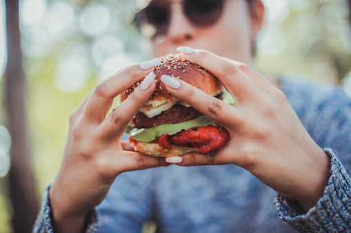 A young woman in a sweater is sitting outside and eating a burger. She is holding it in her hands and enjoying its juiciness as the tomato and lettuce are preparing to fall out.